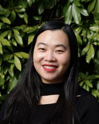 A portrait shot of Christina YZ Chung, who is wearing a black top and red lipstick. She has long black hair, which is worn down. The photo is taken on a sunny day, with green leaves visible in the background.