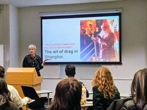 Shelley Pryde stands at a podium presenting during Scholars' Studio. The presentation slide behind them features the title, 'The art of drag in Shanghai,' along with the quote, 'We’re all born naked and the rest is Communism.' A vibrant image of a drag performer is displayed on the slide.