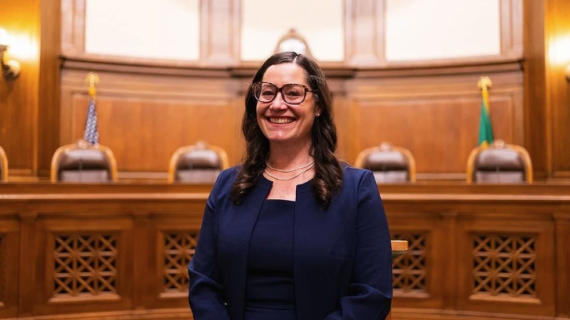 Colleen Melody smiles while seated in a wood-paneled courtroom, wearing glasses and a navy suit. Judicial chairs and the U.S. and Washington State flags appear in the background.
