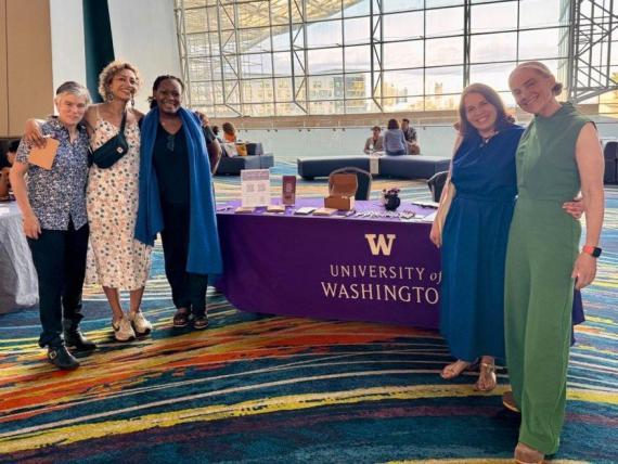 Five GWSS faculty, alumni, and friends stand smiling beside a University of Washington table at the NWSA conference.