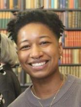 Headshot of Bobbi Kindred smiling at the camera. They have a short afro, and are wearing a thin gold chain and a grey sweater.