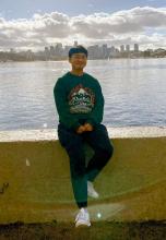 Full body image of Kenji Fujinaka sitting on a cement wall with the Seattle skyline in the background, wearing a green sweatshirt, black running pants, and white gym shoes and smiling at the camera 