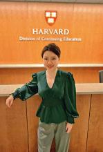Upper body shot of Alva He wearing a green blouse, khaki pants, and her hair in a bun. She is standing in front of a desk with the emblem for Harvard Division of Continuing Education behind her.