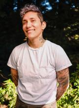 fabian romero standing in front of the woods wearing a pocket t-shirt, with tattoos on arms and neck, hair shaved on the sides and curly on top
