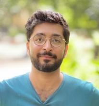Upper body shot of Saad Khan smiling at the camera. He's wearing a light-blue v-neck t-shirt and glasses. The background is blurred greens indicating he may be outside.