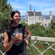 Upper body shot of Jordynn smiling at the camera and giving a thumbs up. She has long brown hair in a ponytail and is wearing a graphic t-shirt. In the background is Germany's famous Neuschwanstein Castle.