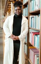 Upper body shot of Ramon wearing a long white jacket with hands clasped in front of him and leaning against a shelf of books. 