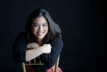 Upper-body portrait of Nina Zafra sitting backward on a chair, smiling broadly with her arms resting across the chair back against a black background. 
