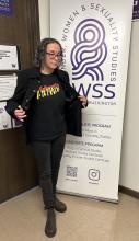 Maria Breaux standing beside a GWSS banner, opening her jacket to show a ‘Vulveeta’ T-shirt.