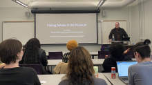 Eric Villiers standing behind a podium in front of a classroom of students. On the screen behind him, the title of his presentation, "Falling Asleep in the Museum"