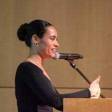 Chloë Bass speaks into a microphone at a podium, shown in profile and gesturing with one hand during her lecture.