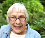 Photo of Carol Allen, an old white woman with ear-length white hair, wearing glasses, a blue button-up over a white t-shirt, with green foliage in the background