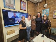 A group of individuals stand shoulder to shoulder smiling in front of a conference room TV with additional individuals on the video call smiling