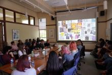 A group of students and faculty sit around a table and in chairs along the wall looking toward a projector screen as a presentation is being shown