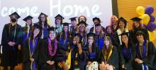 Students of the Class of 2026 stands and crouches for a group photo wearing their black graduation cap and gown in front of a projector screen