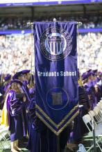 A UW Graduate School banner being held by someone at a graduation ceremony. The person holding the banner is hidden behind it, and in the background you can see a large crowd; the people closer to the camera are wearing purple caps and gowns.