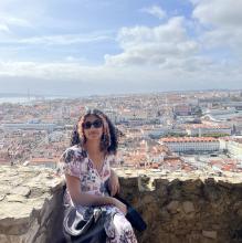 Sana Shetty sitting on a rock wall with the skyline of Lisbon, Portugal in the background. 
