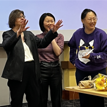 Nancy Kenney and two of her teaching assistants stand at the head of the classroom laughing. In front of them is a table with a bouquet of flowers