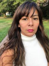 Upper body shot of Iris Viveros Avendano outside with grass and trees behind her. She has long wavy brown hair with long side-swept bangs. She is wearing a white sleeveless turtleneck and pink lipstick. 