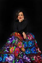 Martha Gonzalez seated against a dark background, wearing a vibrant multicolored skirt with embroidered floral patterns and a black long-sleeved top, resting her chin on her hand with a thoughtful expression.