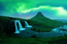 The Northern Lights glow in vibrant green over Kirkjufell Mountain in Iceland. A nearby waterfall cascades into a river, surrounded by lush greenery, with the iconic mountain peak rising in the background under the aurora-lit sky.