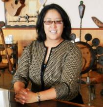 Upper body shot of Regina Yung Lee smiling at the camera, wearing tinted glasses and a black and brown patterned top. She has her hand clasped on the desk in front of her and in the background are a variety of musical instruments.  