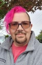 Headshot of Tylir wearing a fleece pullover on top of a pink t-shirt which matches their left-side swept bright pink hair. They are wearing glasses with grey sky and a few trees in the background.