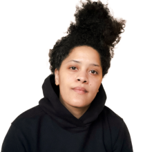 Upper body shot of a Black woman wearing a black hoodie and her afro in a ponytail. She's looking directly at the camera and has a neutral expression.