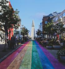 A vibrant rainbow-painted street in Reykjavik leads toward a tall, unique church with a pointed steeple under a clear blue sky. On either side of the street, people enjoy outdoor cafes and shops, with trees and colorful buildings lining the path.