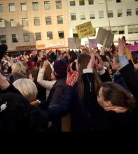 A crowd of people with raised hands protest in a public square, holding signs, including one in Icelandic. The background shows a building with an "American Bar" sign.