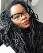 An upper body photo of professor Bettina Judd, a black woman with long braids and her head tilted to the left, wearing black framed glasses and a black top
