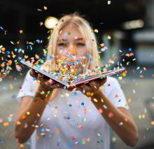 Woman with blonde hair and wearing a plain white t-shirt is holding an open book with multi-colored confetti exploding out of it.