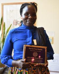 Harriet Dumba, a dark-skinned black woman with long braids pulled back in a ponytail, wearing a blue blouse and patterned skirt, holding her 2022 GWSS Alumni Award plaque and smiling at the camera.