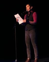 Photo by Ian Johnston/Dangerpants Photography fabian, indigenous two-spirit person, wearing a red dress shirt with a tie and vest, holding a paper while speaking onto a microphone.