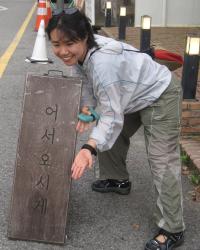 Minjoo Jeong crouches beside a wooden sign with Korean text, smiling and gesturing toward it on a city street.
