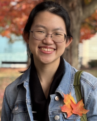 Headshot of young woman with glasses with fall foliage in the background and leaves pinned to her jacket