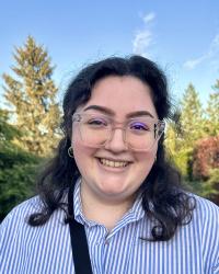 Photograph of Yasmine Gomez in a blue and white striped shirt. She has black curly hair. The background is full of pine trees.