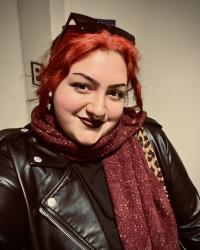 Photograph of Yasmine Gomez. Her hair is red and she is wearing a black jacket.
