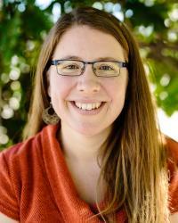 Upper-body photo of Amy Piedalue wearing a bright orange sweater and glasses, with long, straight brown hair.