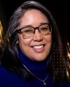 Headshot of Regina Yung Lee smiling. She has long black hair with grey streaks, square framed glasses, and is wearing a royal blue turtleneck sweater with a simple necklace on top. The background is black with gold rays.  