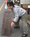 Minjoo Jeong crouches beside a wooden sign with Korean text, smiling and gesturing toward it on a city street.