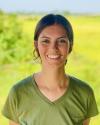 Nastasia is smiling at the camera, standing against a light green backdrop and wearing an olive green t-shirt.