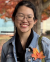 Headshot of young woman with glasses with fall foliage in the background and leaves pinned to her jacket
