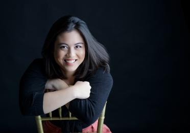 Upper-body portrait of Nina Zafra sitting backward on a chair, smiling broadly with her arms resting across the chair back against a black background. 