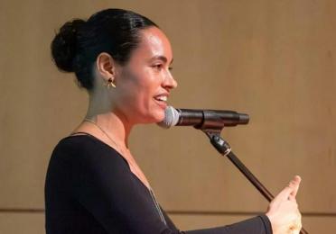 Chloë Bass speaks into a microphone at a podium, shown in profile and gesturing with one hand during her lecture.
