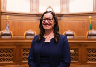 Colleen Melody smiles while seated in a wood-paneled courtroom, wearing glasses and a navy suit. Judicial chairs and the U.S. and Washington State flags appear in the background.
