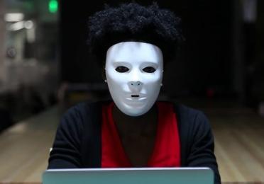 A person wearing a white expressionless mask sits at a laptop in a dimly lit room. They have short curly hair and are dressed in a red shirt and dark cardigan, creating a mysterious, unsettling atmosphere.