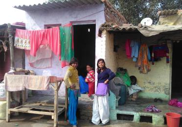 Three young women stand in front of a modest rural home with colorful clothing hung on a clothesline. 