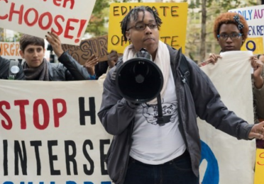 A person with a megaphone standing in front of a group of protesters who are holding signs with messages supporting intersex children's rights. 