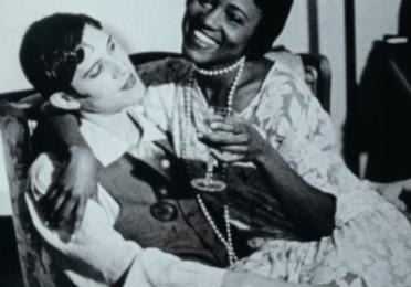Black-and-white film still of two women seated closely together on a couch, smiling and holding drinks, with one woman’s arm wrapped around the other.